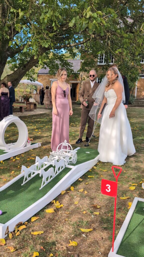 Bride and bridesmaid playing the horse-and-carriage hole at Crockwell Farm, with the ironstone farmhouse behind