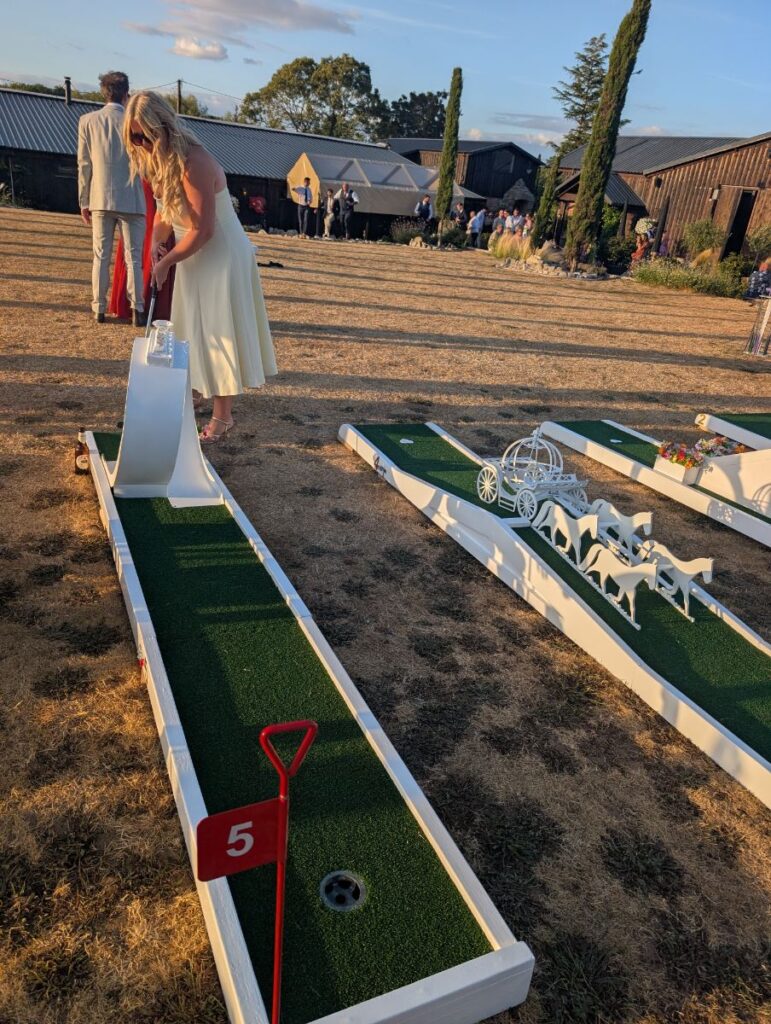 Wedding guest putting on hole 5 of the crazy golf course at The Willow Marsh Farm, with the barns and cypress trees behind